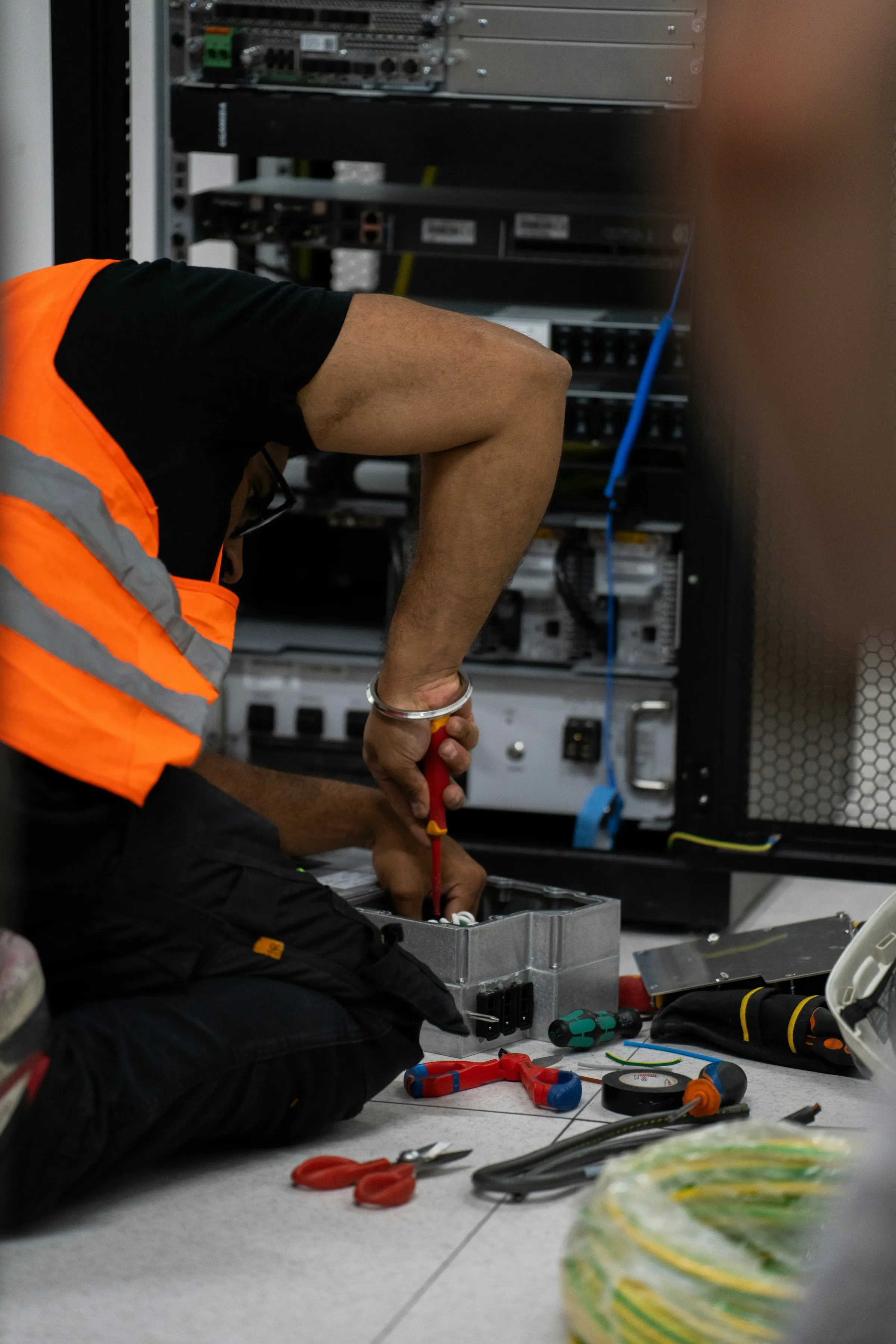 A technician in a hi-vis vest working at a network rack with tools laid out on the floor